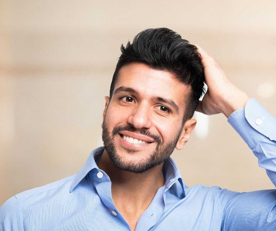 Smiling man with a beard in a blue shirt, touching his hair, against a neutral background.